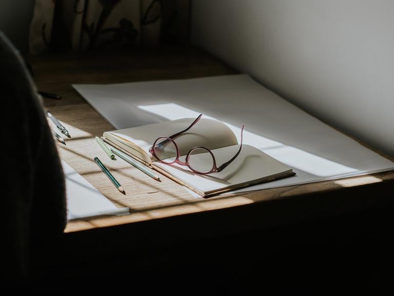 Eine Brille, die auf einem leeren Notizbuch liegt. Stifte umgeben das Buch, und Sonnenlicht fällt durch das Fenster und wirft Sonnenstrahlen auf den Schreibtisch. Eine Brille, die auf einem leeren Notizbuch liegt. Stifte umgeben das Buch, und Sonnenlicht fällt durch das Fenster und wirft Sonnenstrahlen auf den Schreibtisch.