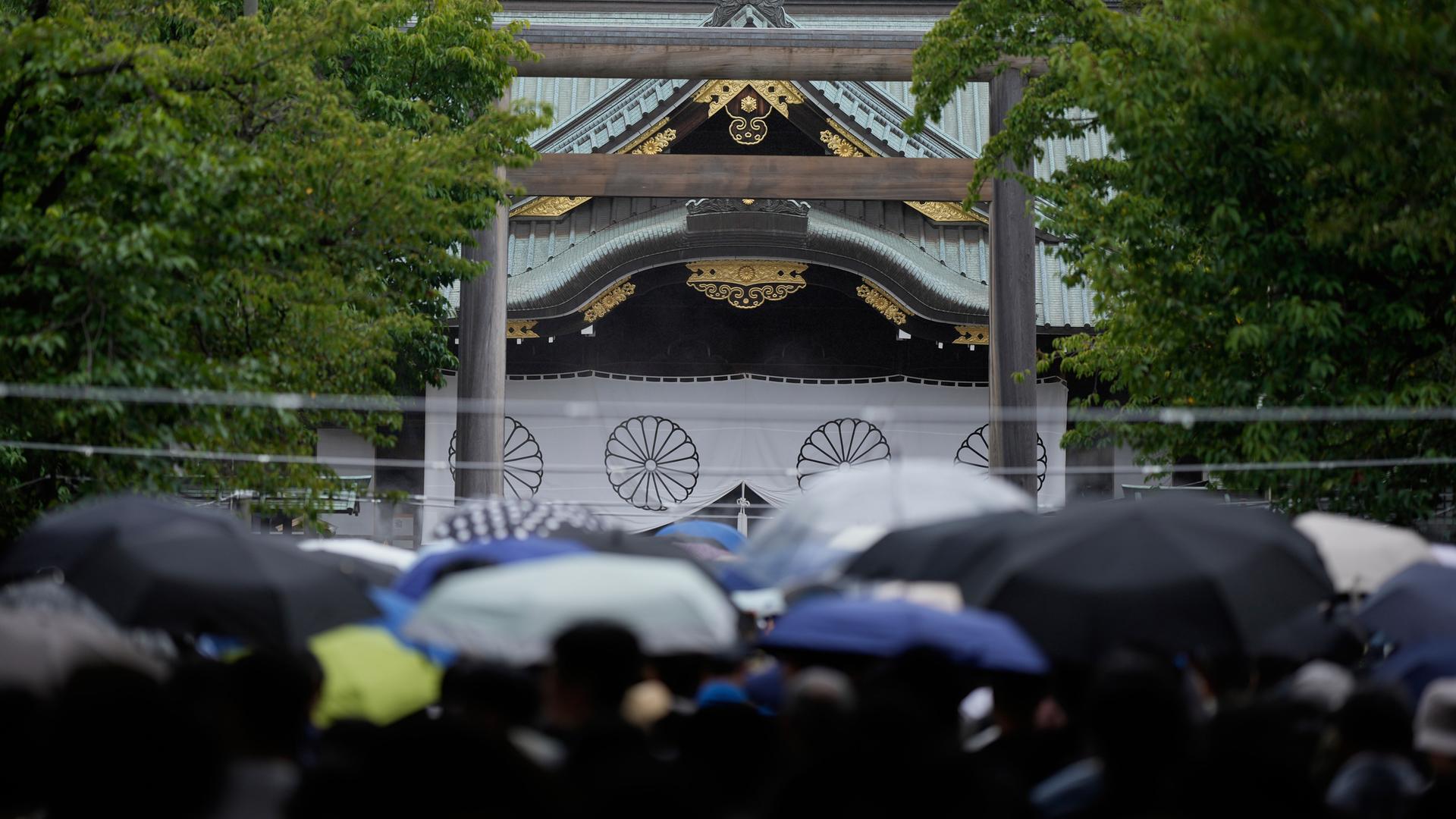 Eine Menschenansammlung mit Regenschirmen steht vor einem japanischen Schrein. Eine Menschenansammlung mit Regenschirmen steht vor einem japanischen Schrein.