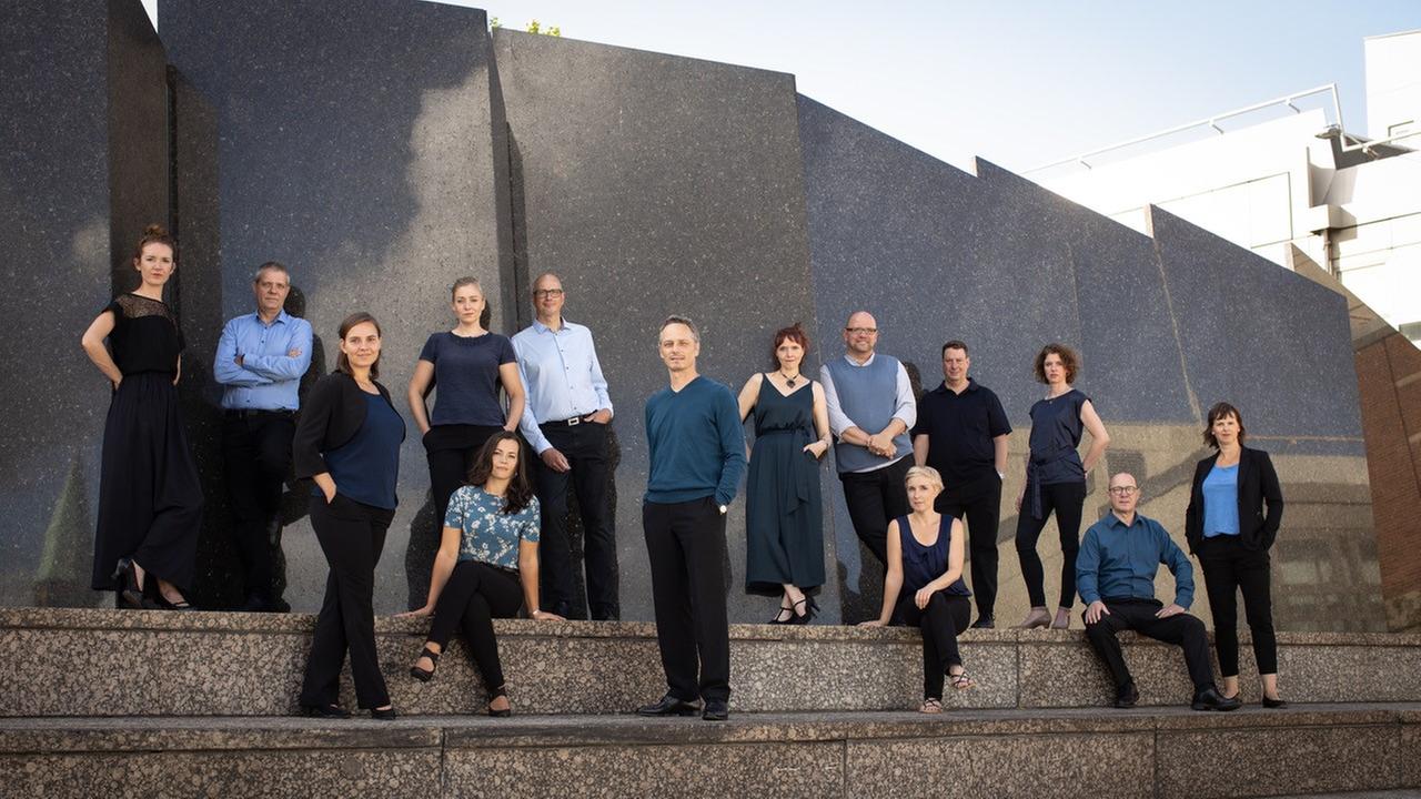 Das Bild zeigt den Vocalconsort mit insgesamt 14 Personen, das vor einer Steinwand auf Treppenstufen stehend und im sitzen posiert. Das Bild zeigt den Vocalconsort mit insgesamt 14 Personen, das vor einer Steinwand auf Treppenstufen stehend und im sitzen posiert.