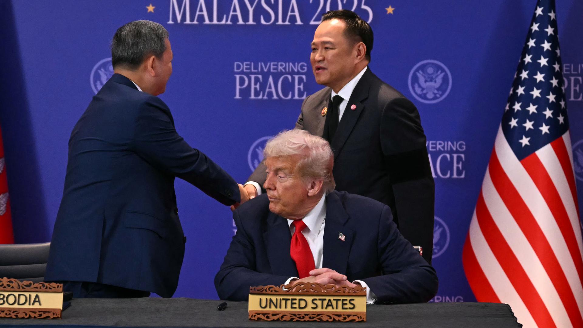 Zwei Männer in Anzügen schütteln sich stehend die Hände. Im Vordergrund sitzt ein weiterer Mann in Anzug und roter Krawatte vor einem Tischaufstellerchen. Darauf steht: United States. Zwei Männer in Anzügen schütteln sich stehend die Hände. Im Vordergrund sitzt ein weiterer Mann in Anzug und roter Krawatte vor einem Tischaufstellerchen. Darauf steht: United States.