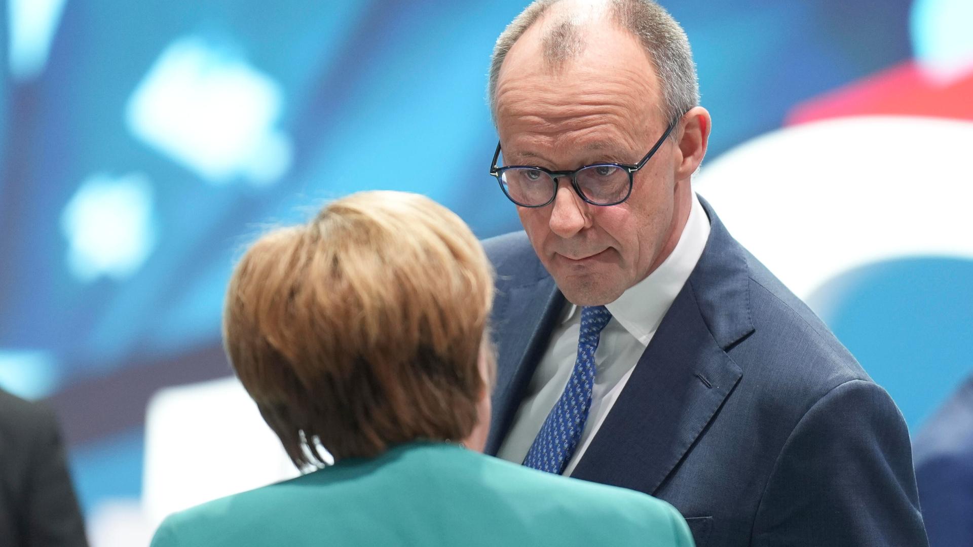 Friedrich Merz im Gespräch mit Angela Merkel. Merkel steht mit dem Rücken zur Kamera, Merz schaut sie mit konzentriertem Gesichtsausdruck an.