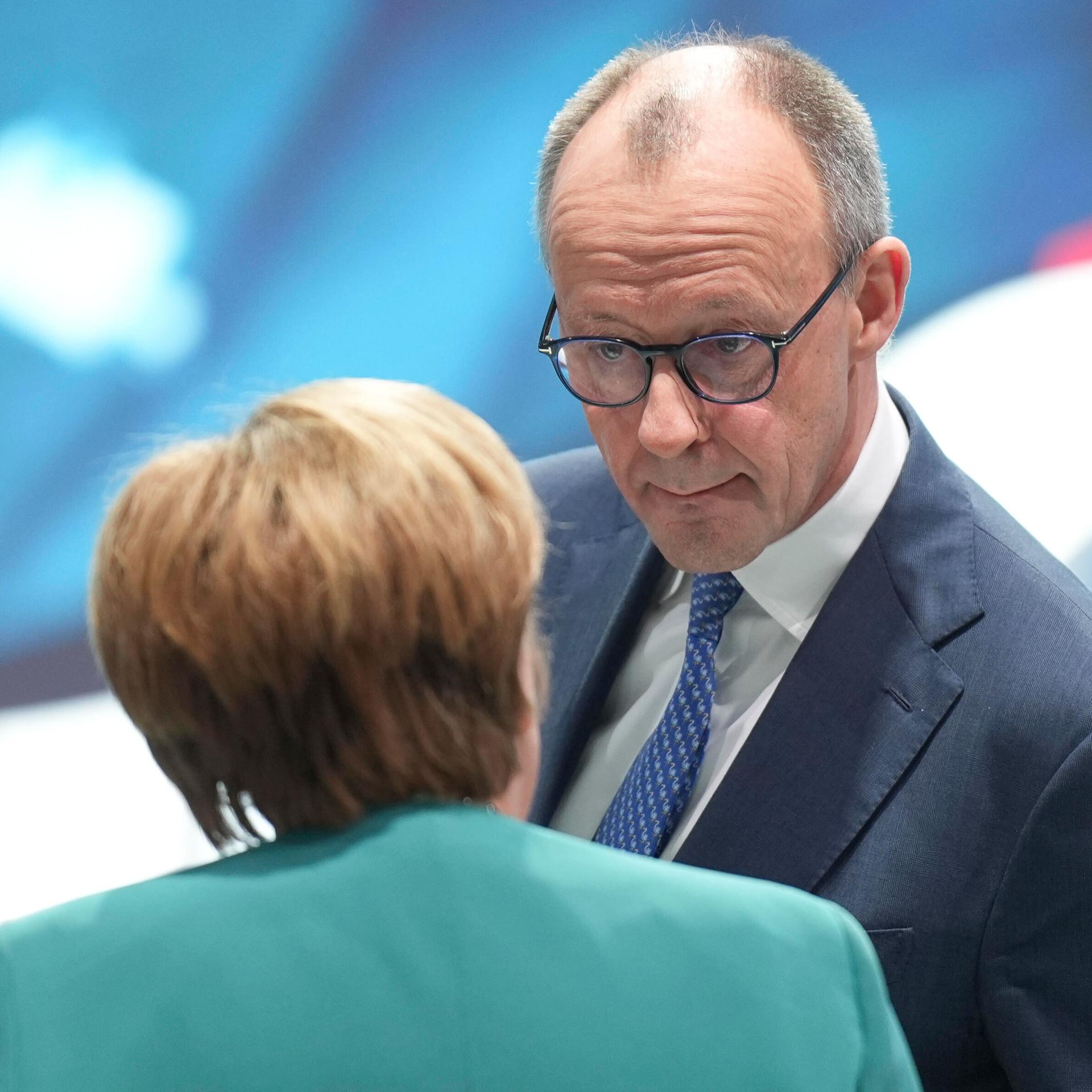 Das Foto zeigt Friedrich Merz im Gespräch mit Angela Merkel. Merkel steht mit dem Rücken zur Kamera, Merz schaut sie mit konzentriertem Gesichtsausdruck an.