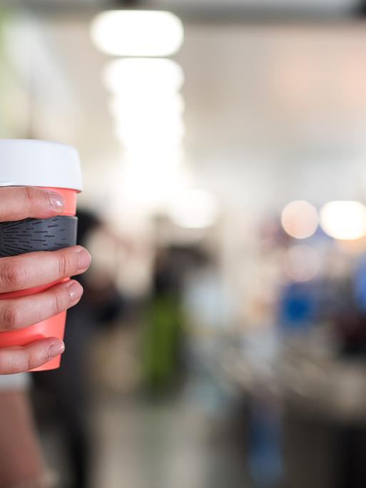 Eine Frau hält einen Mehrweg-Becher für Kaffee in der Hand. Es ist nur ihre Hand zu sehen, dahinter ein Cafe.