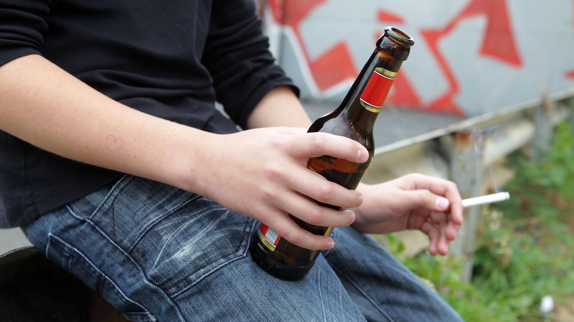 Man sieht den Körper eines Jugendlichen. Er hat eine Flasche Bier und eine Zigarette in den Händen.