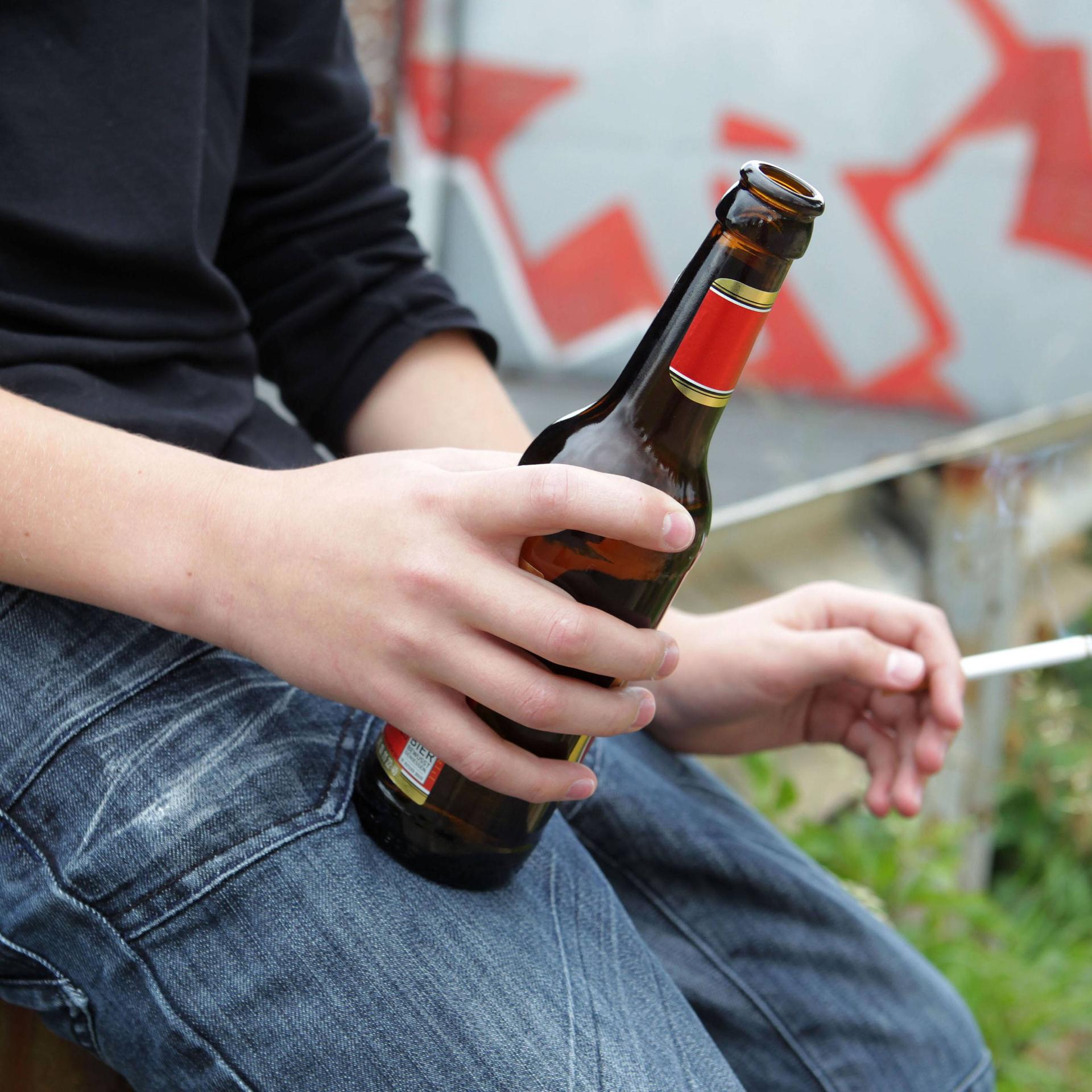 Man sieht den Körper eines Jugendlichen. Er hat eine Flasche Bier und eine Zigarette in den Händen.