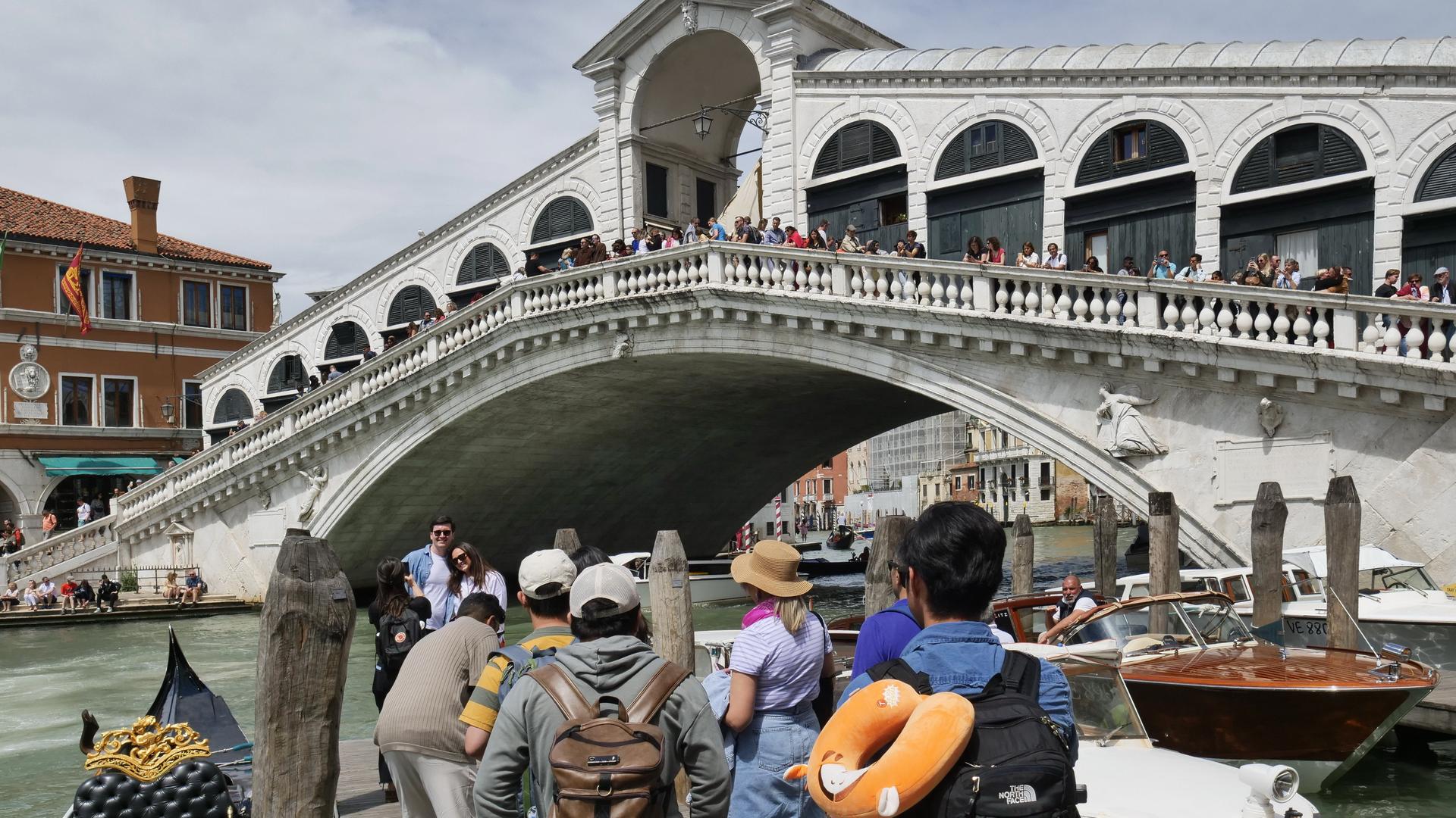 Eine Gruppe von Besuchern steht in der Stadt Venedig am Wasser. Im Hintergrund ist eine Brücke mit vielen Menschen zu sehen. 