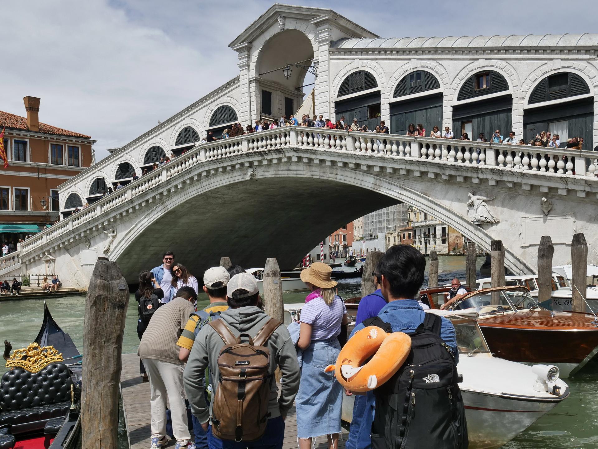 Eine Gruppe von Touristen steht in Venedig am Wasser. Im Hintergrund ist eine von Menschen überfüllte Brücke zu sehen.