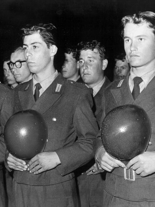 Historisches Schwarzweißfoto von Bundeswehrrekruten während der feierlichen Verpflichtung. Sie stehen aufgereiht nebeneinander und halten ihre Helme vor der Brust.
