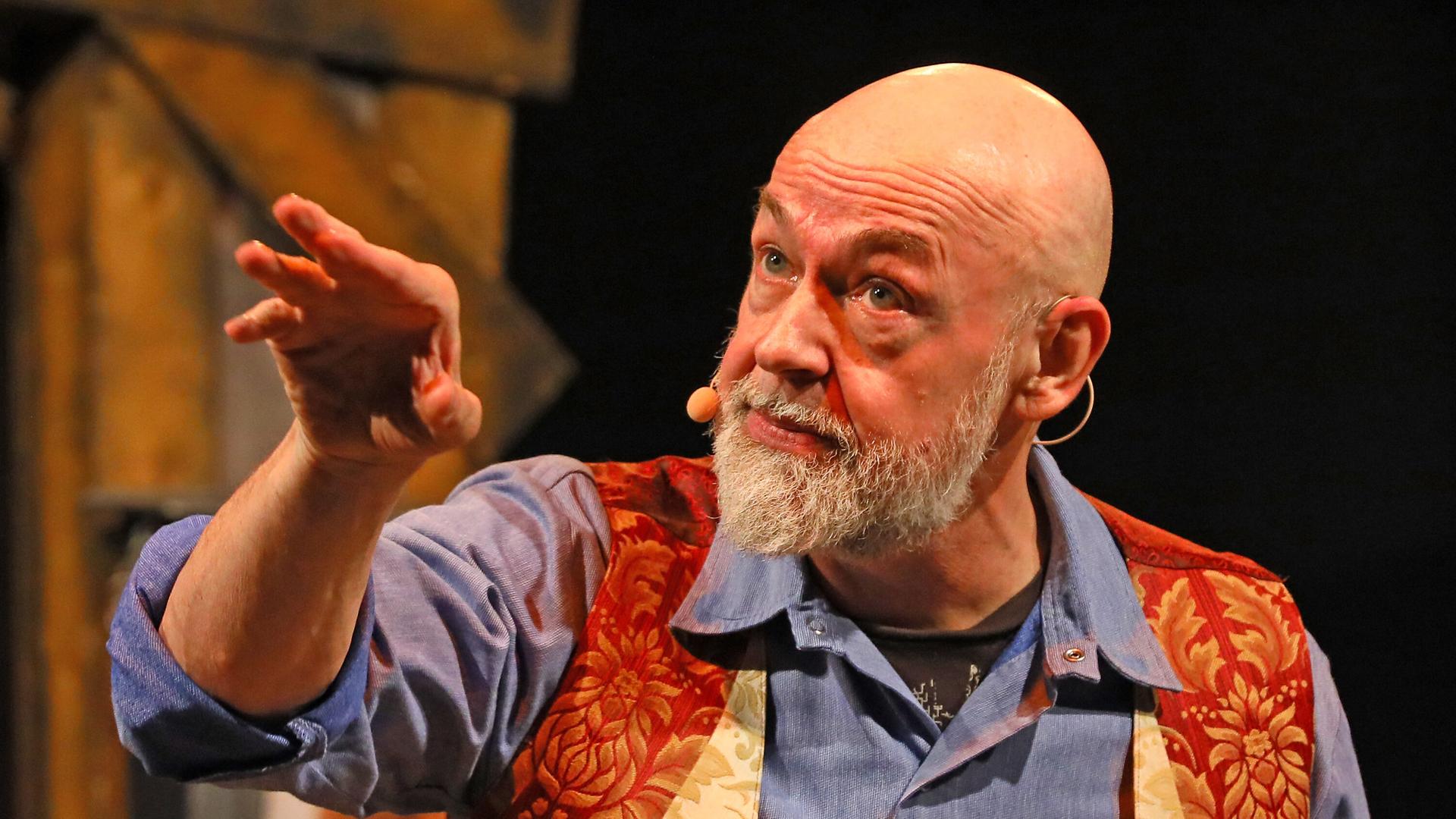 Gerd Knebel zeigt bei einem Auftritt mit dem Arm zur Seite. Er trägt ein blaues Hemd und eine rot-goldene Weste.