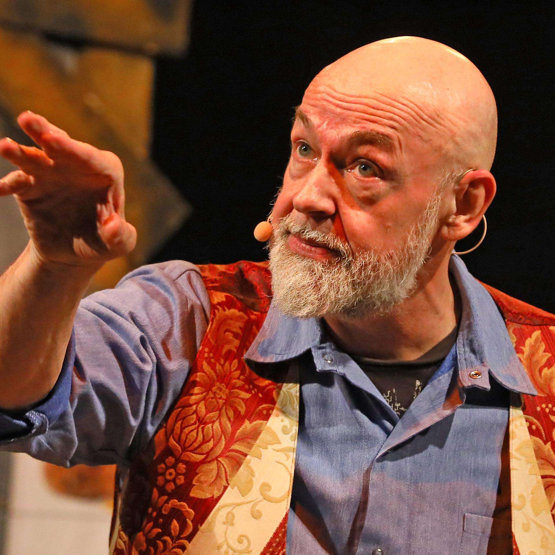 Gerd Knebel zeigt bei einem Auftritt mit dem Arm zur Seite. Er trägt ein blaues Hemd und eine rot-goldene Weste.