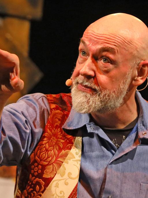 Gerd Knebel zeigt bei einem Auftritt mit dem Arm zur Seite. Er trägt ein blaues Hemd und eine rot-goldene Weste.