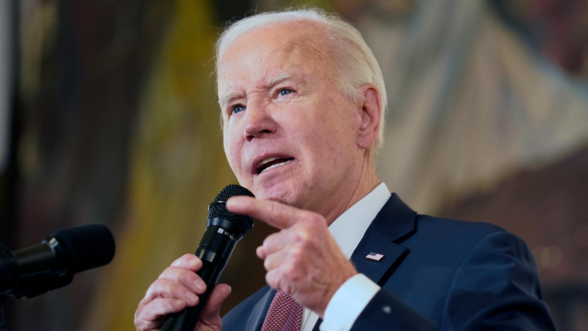 Biden spricht vor einem bräunlichen Hintergrund in ein Mikrofon und zeigt dabei mit der linken Zeigefinger nach vorne.