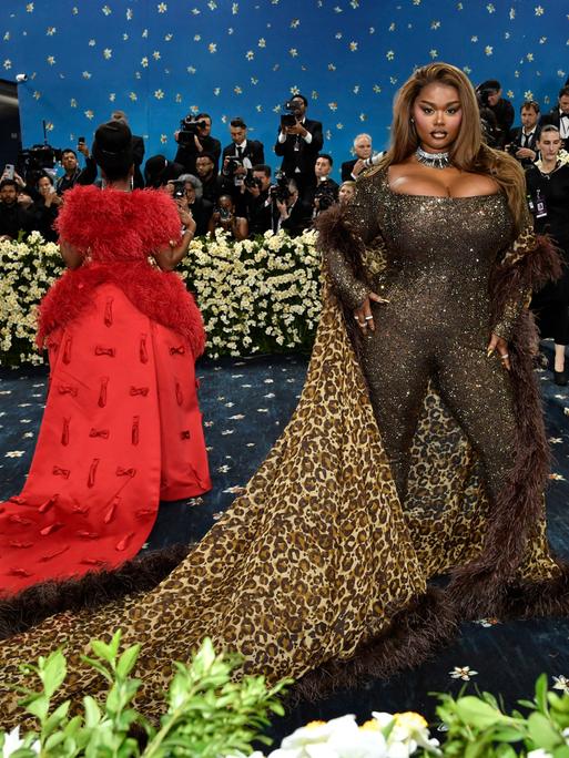 Precious Lee zeigt sich bei der Met Gala den Fotografen. Sie trägt einen engen Glitzeranzug und einen Umhang mit Leoparden-Muster. Neben ihr eine weitere Frau mit auslander roter Schleppe von hinten. 
