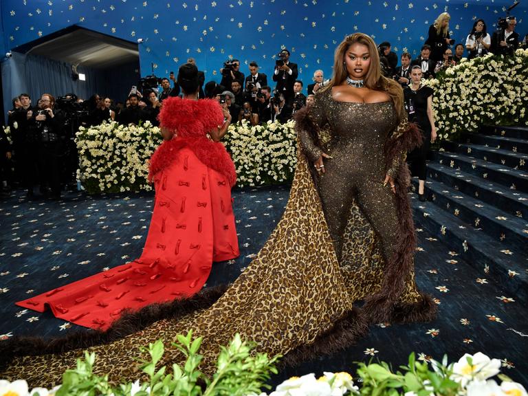 Precious Lee zeigt sich bei der Met Gala den Fotografen. Sie trägt einen engen Glitzeranzug und einen Umhang mit Leoparden-Muster. Neben ihr eine weitere Frau mit auslander roter Schleppe von hinten. 