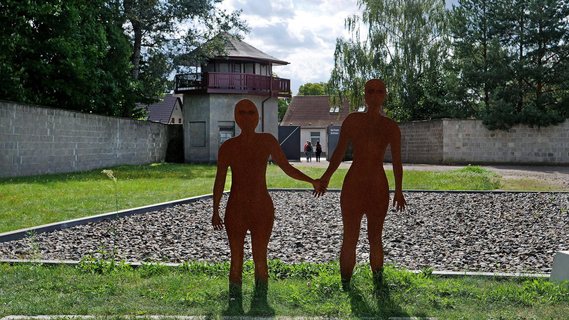 Gedenkstätte im ehemaligen Konzentrationslager Sachsenhausen in Oranienburg bei Berlin. Die Plastiken zweier Menschen-Silhouetten stehen Hand in Hand auf einer Rasenfläche. Im Hintergrund ist ein Wachturm.