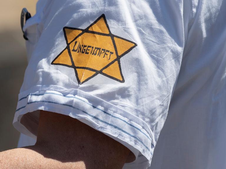 "Ungeimpft" steht auf einem nachgebildeten "Gelben Stern" am Arm eines Mannes. Dieses Zwangskennzeichen diente in der NS-Zeit dazu, jüdische Bürger zu diskriminieren. 