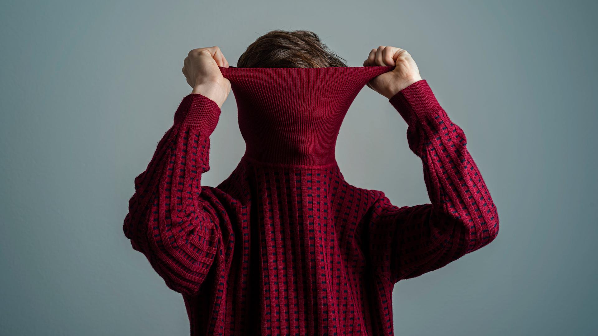 Ein Mann zieht einen weinroten Rollkragen-Pullover so über den Kopf, dass man sein Gesicht nicht mehr erkennen kann.