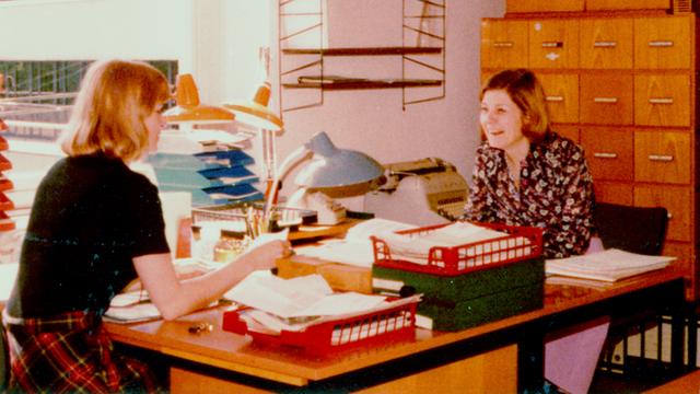 Zwei Frauen sitzen sich in einem Büro an zwei Schreibtischen gegenüber und unterhalten sich