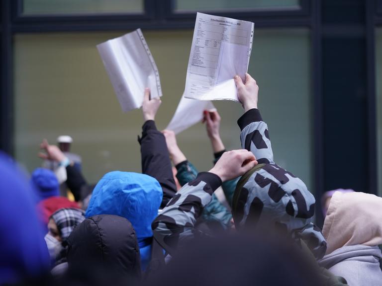 Flüchtlinge halten Formulare in einer Warteschlange vor einer Ausländerbehörde hoch.