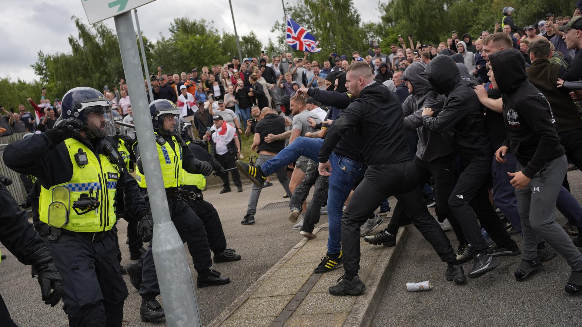 Eine große Gruppe von Demonstranten steht auf einer Straße zwei Polizisten gegenüber. Ein Demonstrant scheint nach einem der Polizisten zu treten.