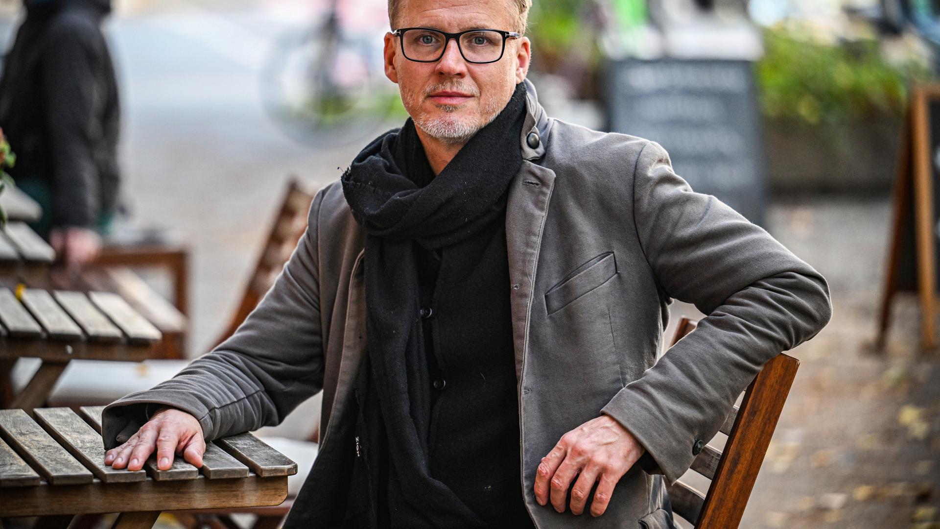 Falk Richter sitzt an einem Tisch in einem Biergarten. Seine Hand liegt auf dem Tisch, er trägt Mantel und Schal sowie eine schwarz umrandete Brille.