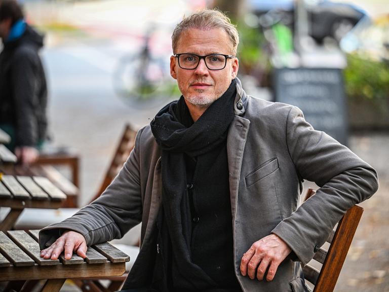 Falk Richter sitzt an einem Tisch in einem Biergarten. Seine Hand liegt auf dem Tisch, er trägt Mantel und Schal sowie eine schwarz umrandete Brille.