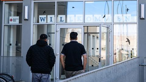 Zwei Asylbewerber gehen in das Gebäude. Sie sind nur von hinten vor einer Glastür zu sehen. 
