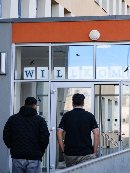 Zwei Asylbewerber gehen in das Gebäude. Sie sind nur von hinten vor einer Glastür zu sehen. 