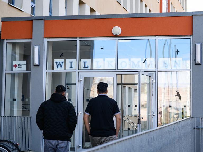 Zwei Asylbewerber gehen in das Gebäude. Sie sind nur von hinten vor einer Glastür zu sehen. 