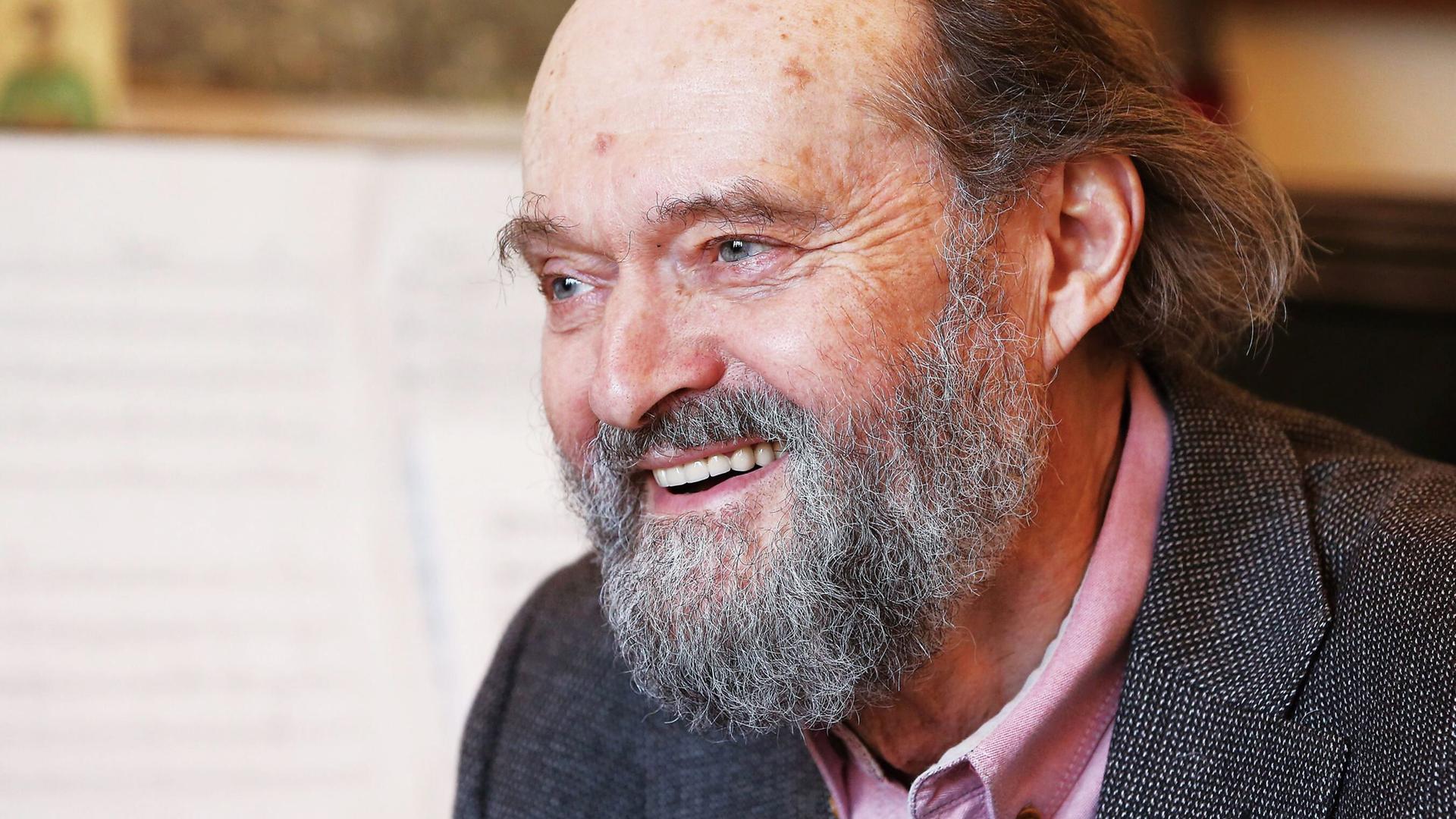 Arvo Pärt sitzt mit Bart lachend vor einem Flügel, auf dem Noten aufgeschlagen sind.