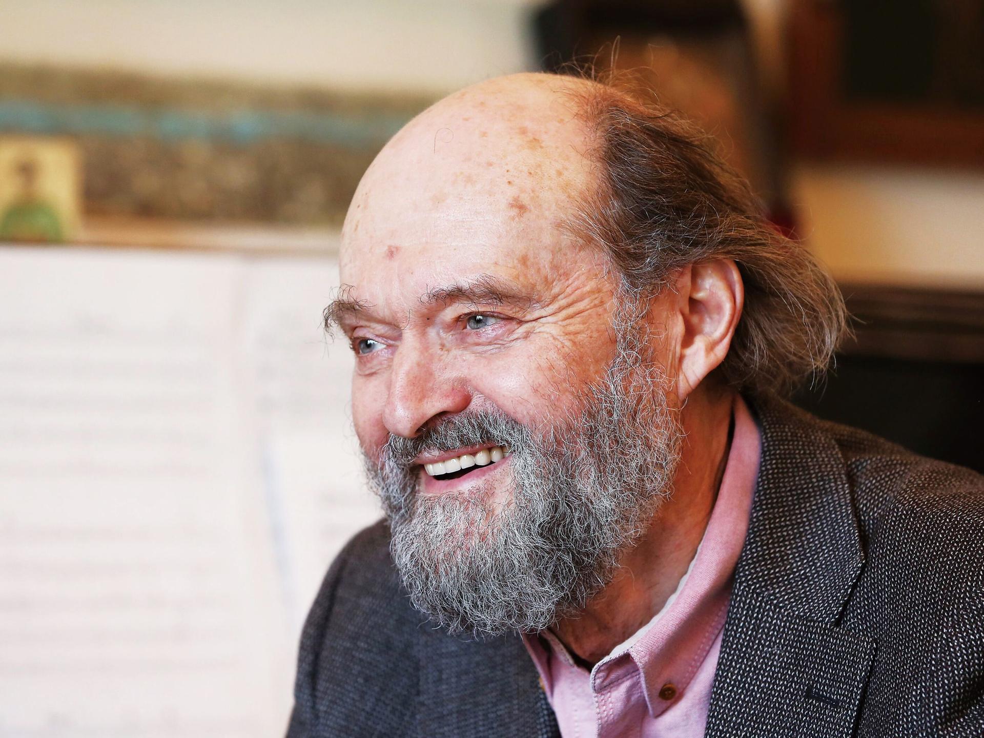 Arvo Pärt sitzt mit Bart lachend vor einem Flügel, auf dem Noten aufgeschlagen sind.