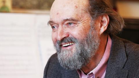 Arvo Pärt sitzt mit Bart lachend vor einem Flügel, auf dem Noten aufgeschlagen sind.