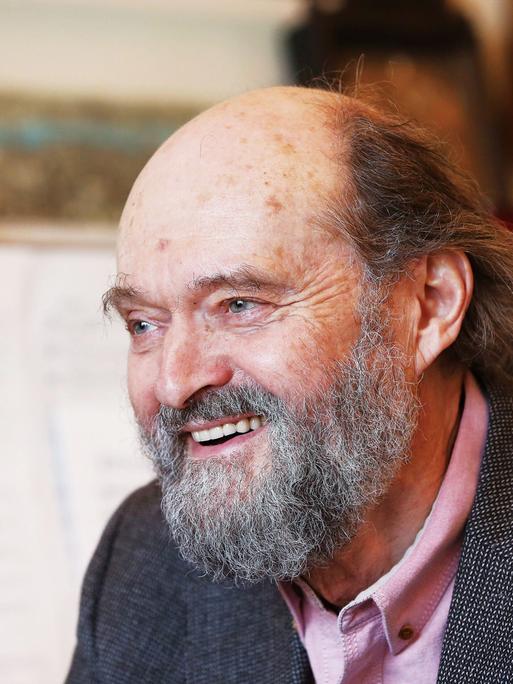 Arvo Pärt sitzt mit Bart lachend vor einem Flügel, auf dem Noten aufgeschlagen sind.