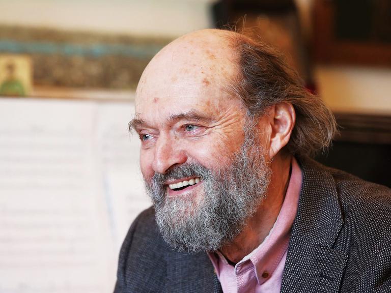 Arvo Pärt sitzt mit Bart lachend vor einem Flügel, auf dem Noten aufgeschlagen sind.