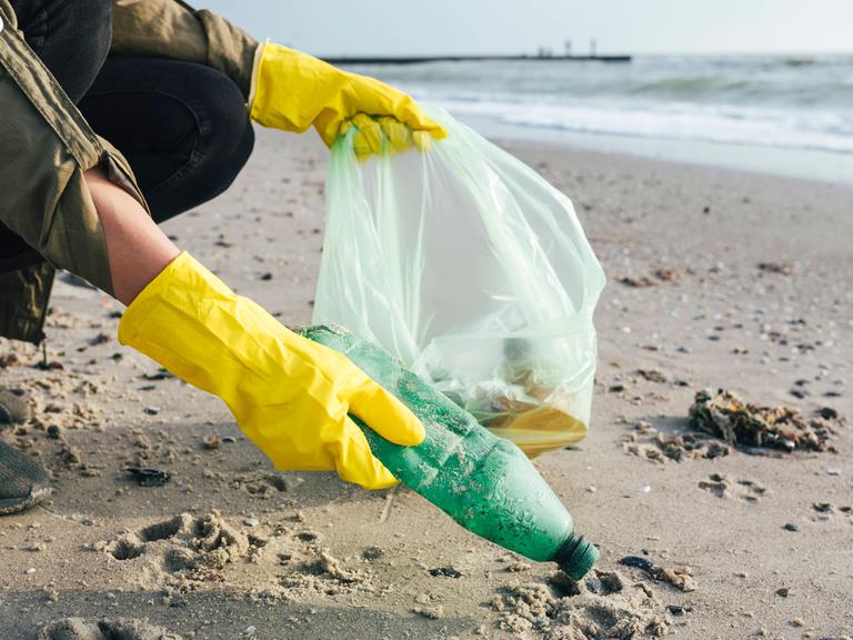 Eine Person sammlt Plastikmüll am Strand. 