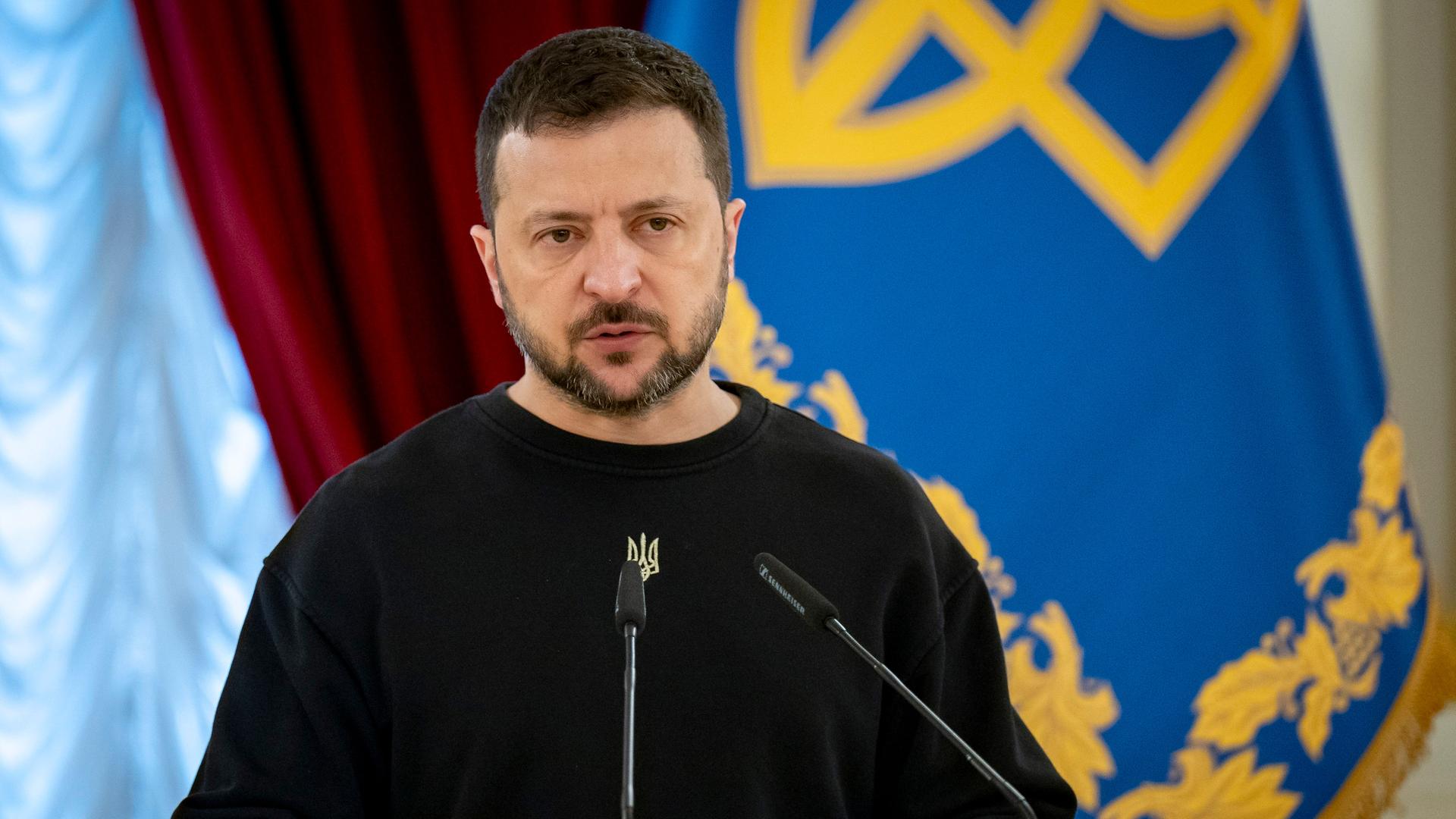 Wolodymyr Selenskyj auf einer Pressekonferenz, im Hintergrund sind verschiedene Flaggen zu sehen.