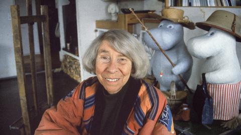 Eine ältere Frau mit grauen Haaren sitzt in einem Atelier. Es ist die Schriftstellerin Tove Jansson. Hinter ihr sind Figuren der Mumins, die Jansson erfunden hat.