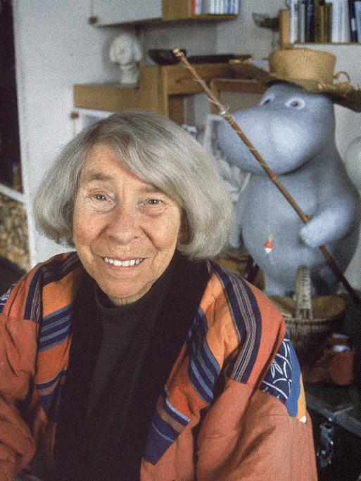 Eine ältere Frau mit grauen Haaren sitzt in einem Atelier. Es ist die Schriftstellerin Tove Jansson. Hinter ihr sind Figuren der Mumins, die Jansson erfunden hat.