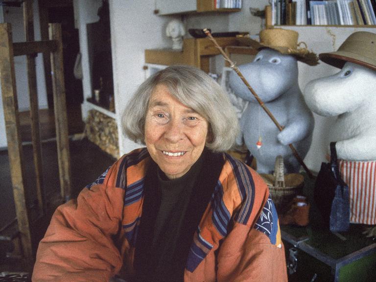 Eine ältere Frau mit grauen Haaren sitzt in einem Atelier. Es ist die Schriftstellerin Tove Jansson. Hinter ihr sind Figuren der Mumins, die Jansson erfunden hat.