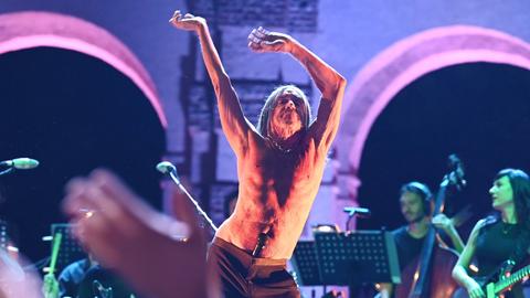 Der 75-jährige Sänger Iggy Pop tanzt mit freiem Oberkörper während eines Konzerts auf der Bühne des Palazzo Te in Mantua. In der Hüfte knickt er seitlich ab, die Hände hat er hoch in der Luft, das Mikrofon steckt im Hosenbund.