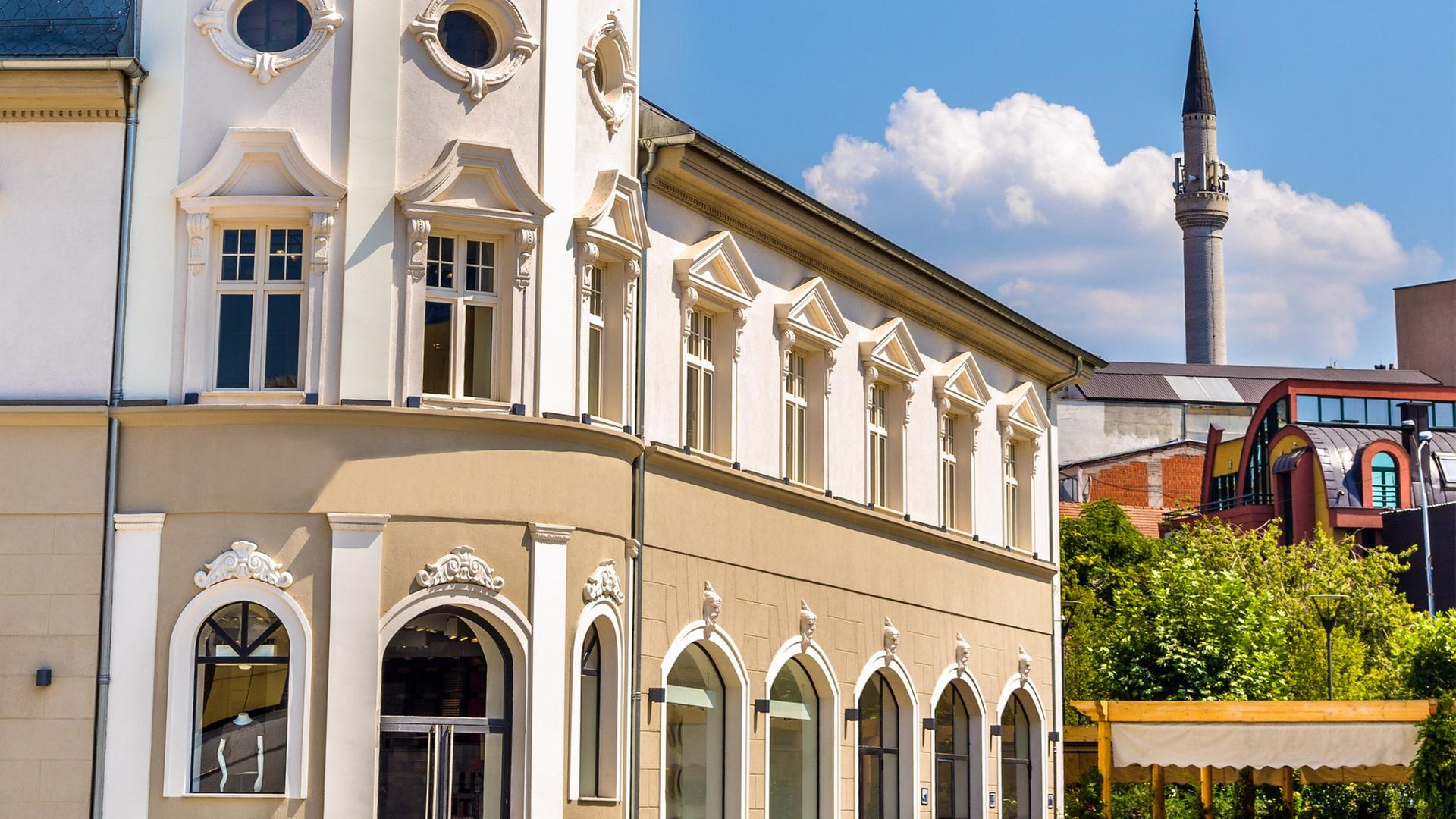 Ein prächtiges Gebäude in Pristina mit Stuck an der Fassade, im Hintergrund eine Terrasse, auf der Menschen sitzen, und weitere Gebäude.