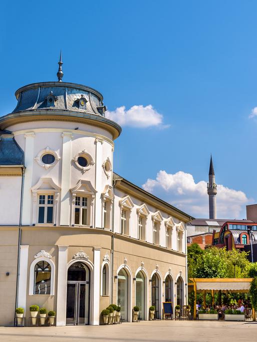 Ein prächtiges Gebäude in Pristina mit Stuck an der Fassade, im Hintergrund eine Terrasse, auf der Menschen sitzen, und weitere Gebäude.