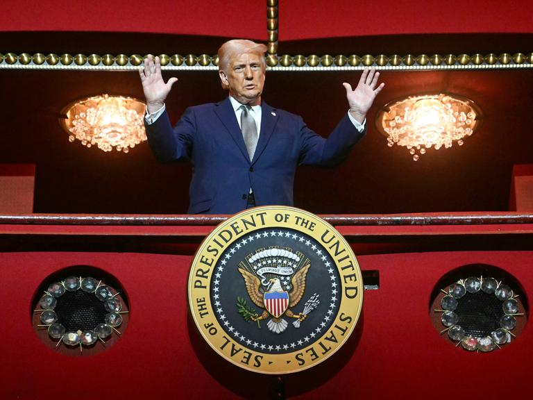 US-Präsident Donald Trump steht mit erhobenen Armen zwischen zwei Kronleuchtern in einer Loge des Kennedy Center in Washington, an der Brüstung ist ein rundes Schild mit dem Präsidentenwappen angebracht: Seal of the President of the United States.