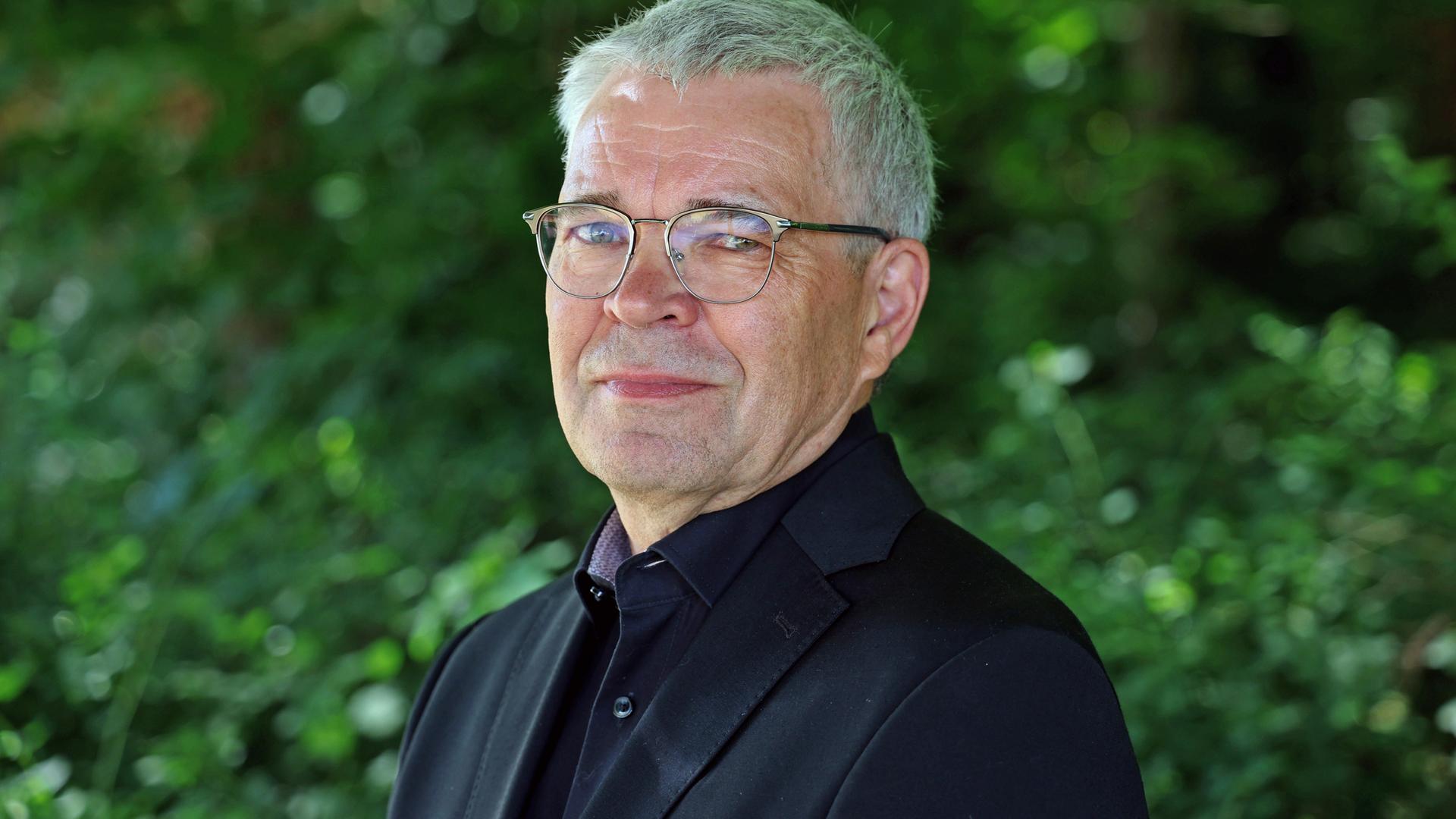 Fred Zack lächelt in die Kamera. Er trägt ein schwarzes Hemd zu schwarzem Anzug und eine silberne Brille. Fred Zack lächelt in die Kamera. Er trägt ein schwarzes Hemd zu schwarzem Anzug und eine silberne Brille.