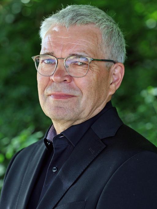 Fred Zack lächelt in die Kamera. Er trägt ein schwarzes Hemd zu schwarzem Anzug und eine silberne Brille.