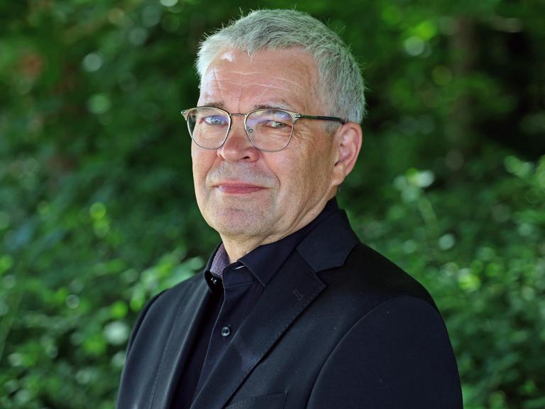 Fred Zack lächelt in die Kamera. Er trägt ein schwarzes Hemd zu schwarzem Anzug und eine silberne Brille.