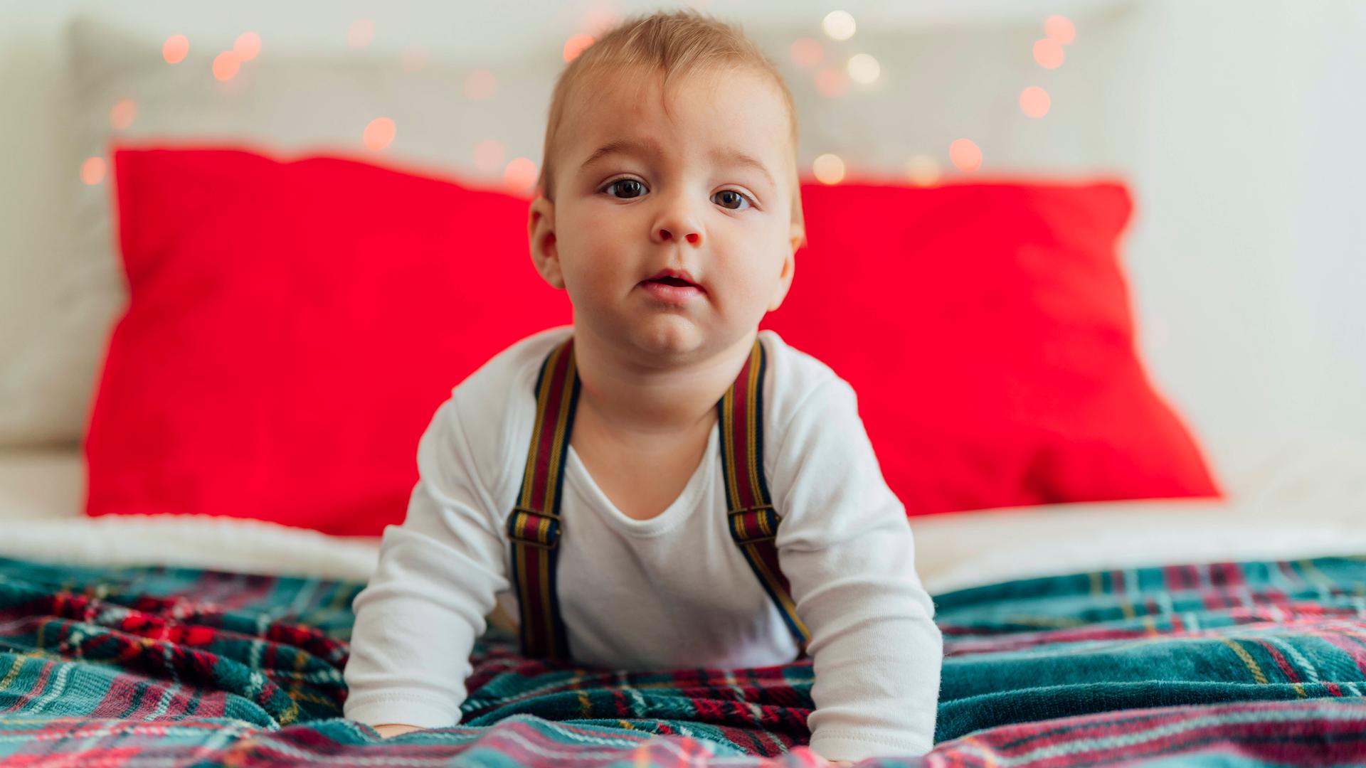 Ein größeres Baby mit Hosenträgern liegt auf einem weihnachtlich geschmückten Bett und stemmt Kopf und Oberkörper in die Höhe und blickt in die Kamera.