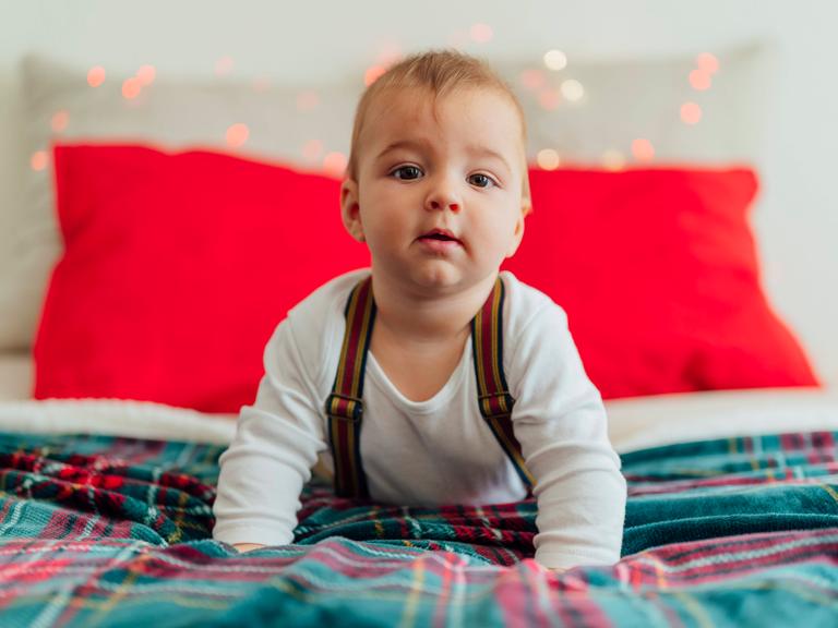Ein größeres Baby mit Hosenträgern liegt auf einem weihnachtlich geschmückten Bett und stemmt Kopf und Oberkörper in die Höhe und blickt in die Kamera.