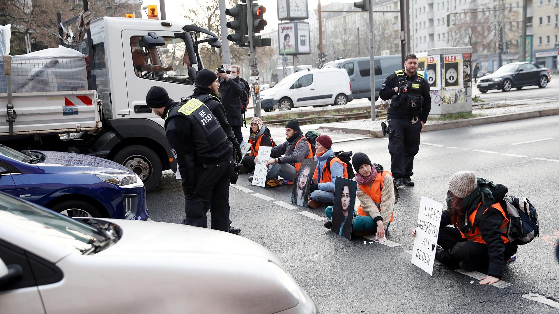 Aktivisten in Warnwesten sitzen in einer Reihe auf der Straße, umgeben von Autos.