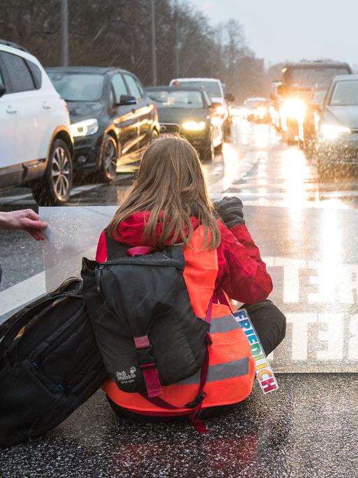 Zwei Klimaaktivisten sitzen mit Warnwesten und einem Transparent auf einer Straße. Vor ihnen stehen viele Autos.
